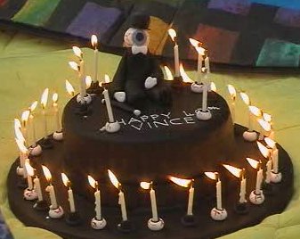 A crudely tophat-shaped cake with an eyeball Resident sitting on top. There are several candles on the cake being held up by tiny eyeballs.